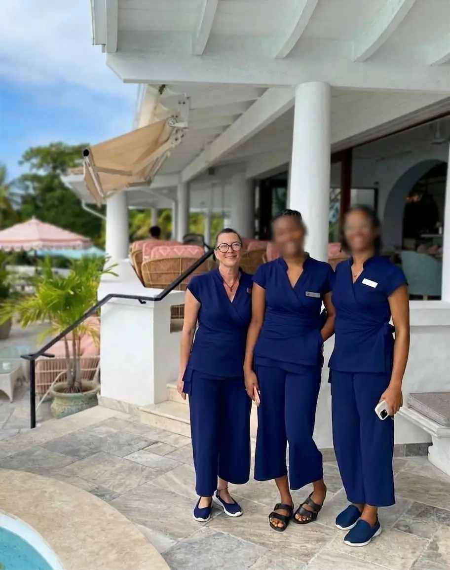 A medium shot of three smiling female massage therapists at the La Samanna spa, standing on the resort's outdoor stone terrace in St Martin. On the left is Virginie, wearing glasses, a navy blue wrap tunic, matching trousers, and blue flat shoes. To her right stand two colleagues with their faces blurred for privacy, wearing the same navy blue uniform. The middle colleague wears black sandals, and the one on the right holds a white smartphone and wears blue trainers. The background features the resort's white architecture, rattan lounge furniture, white pillars, palm trees, and a view of the turquoise sea. The floor is made of grey-blue stone slabs.