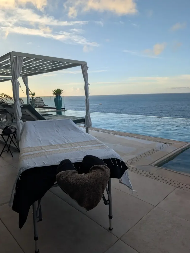 A bespoke massage table setup featuring Uhaina’s signature textured cloth on a luxurious villa terrace in St. Martin, as seen in image PXL_20260212_111024001.MP.jpg. In the midground are private white cabanas, a distinctive turquoise planter, and an infinity pool, all overlooking the vast ocean at golden hour.